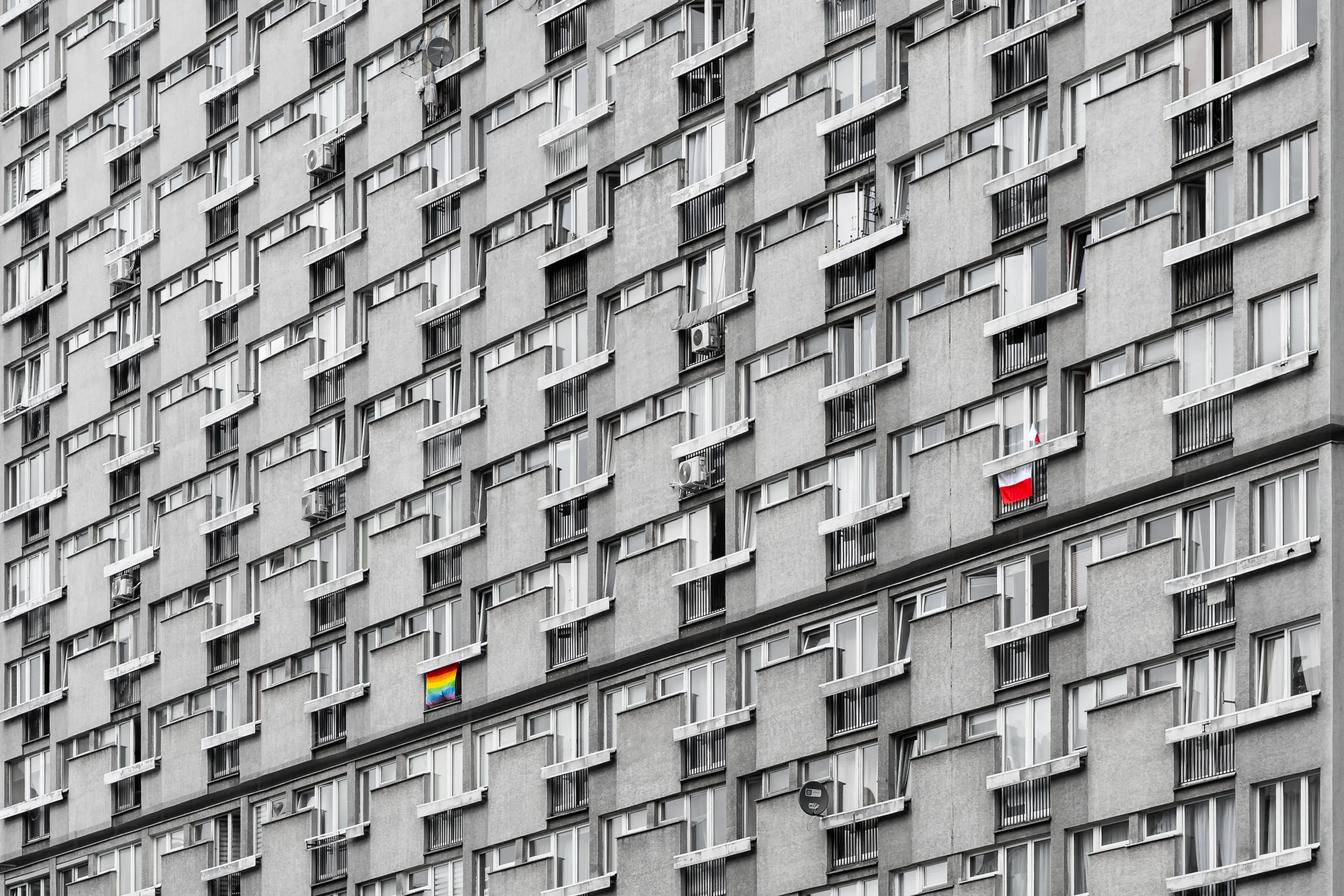 Black-and-white facade of a Warsaw high-rise, its grid of balconies broken by a small rainbow flag.