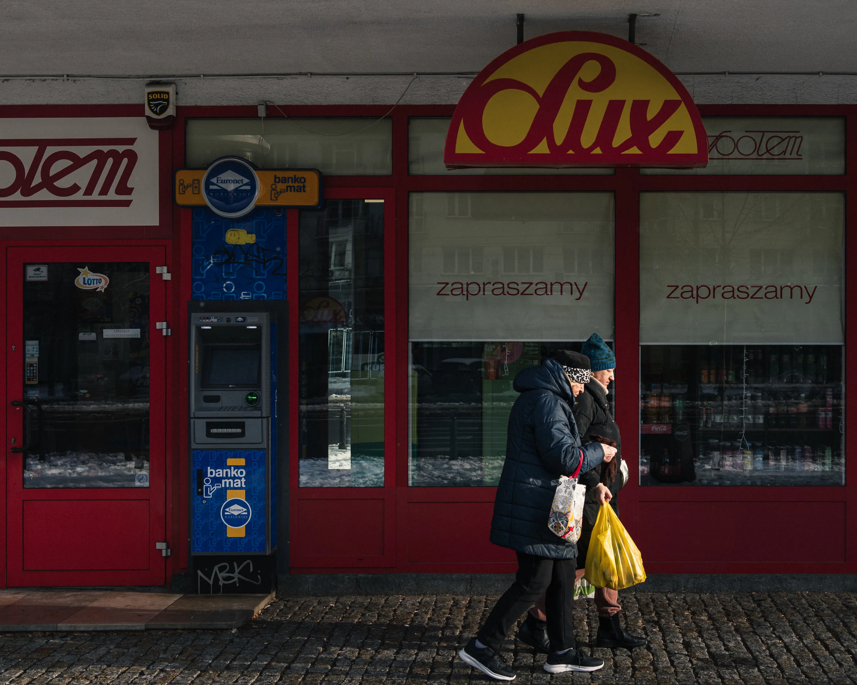 An elderly couple walks past a corner shop's Lux sign and cash machine in soft winter light.