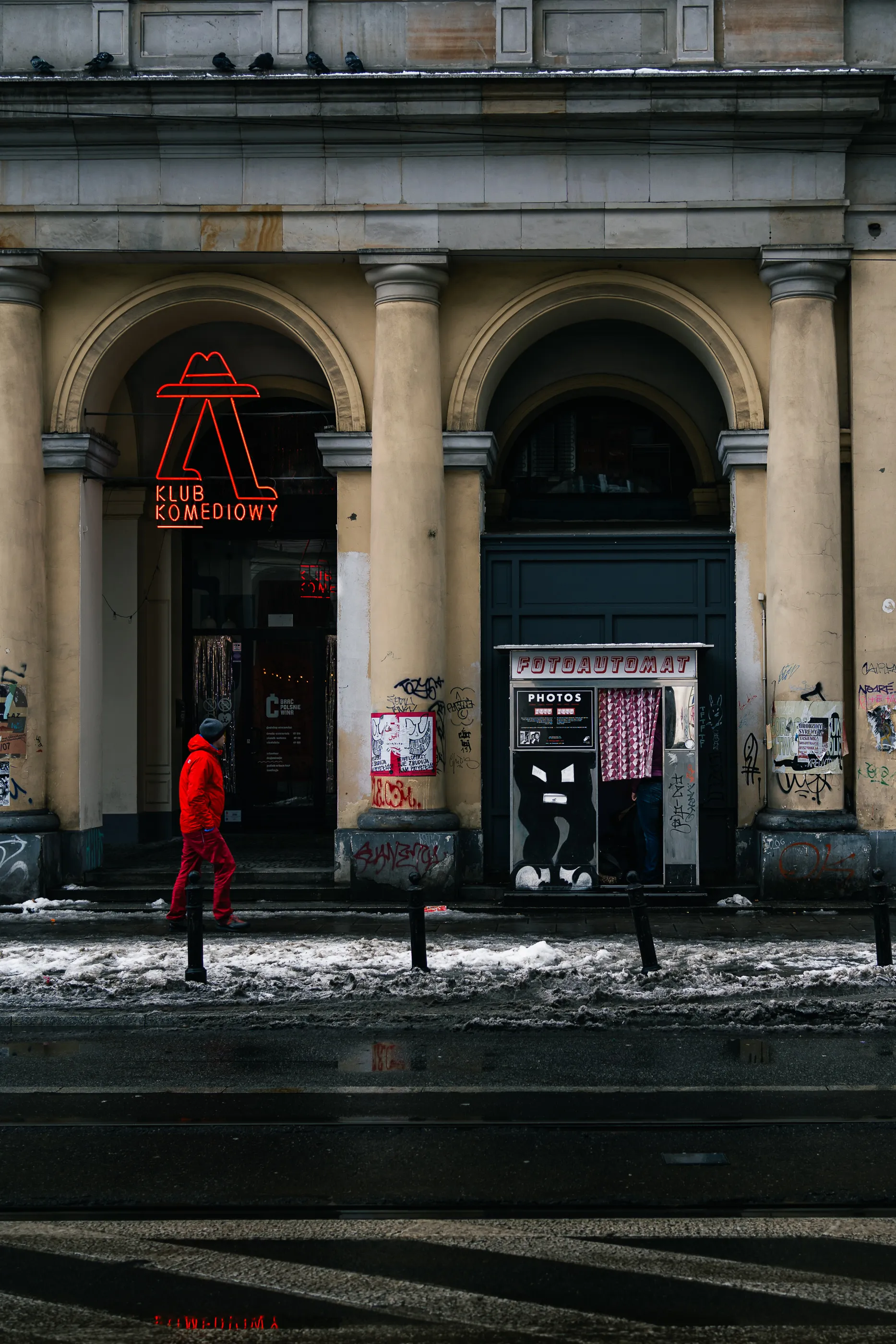 A man in a red jacket walks past a neon comedy-club sign and a photo booth on a snowy Warsaw street.