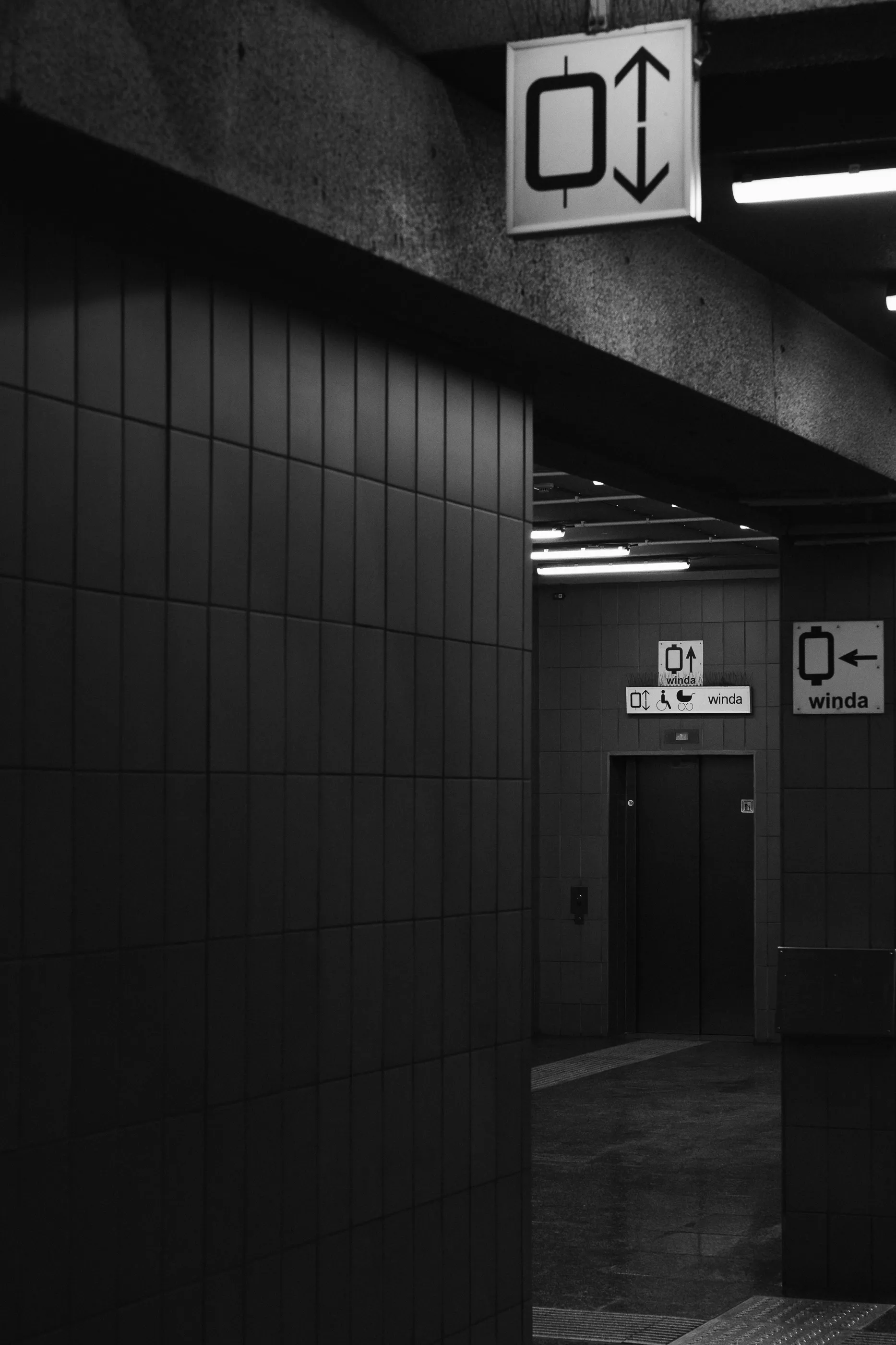Black-and-white elevator signs ("winda") glow above a dim, tiled underground corridor.