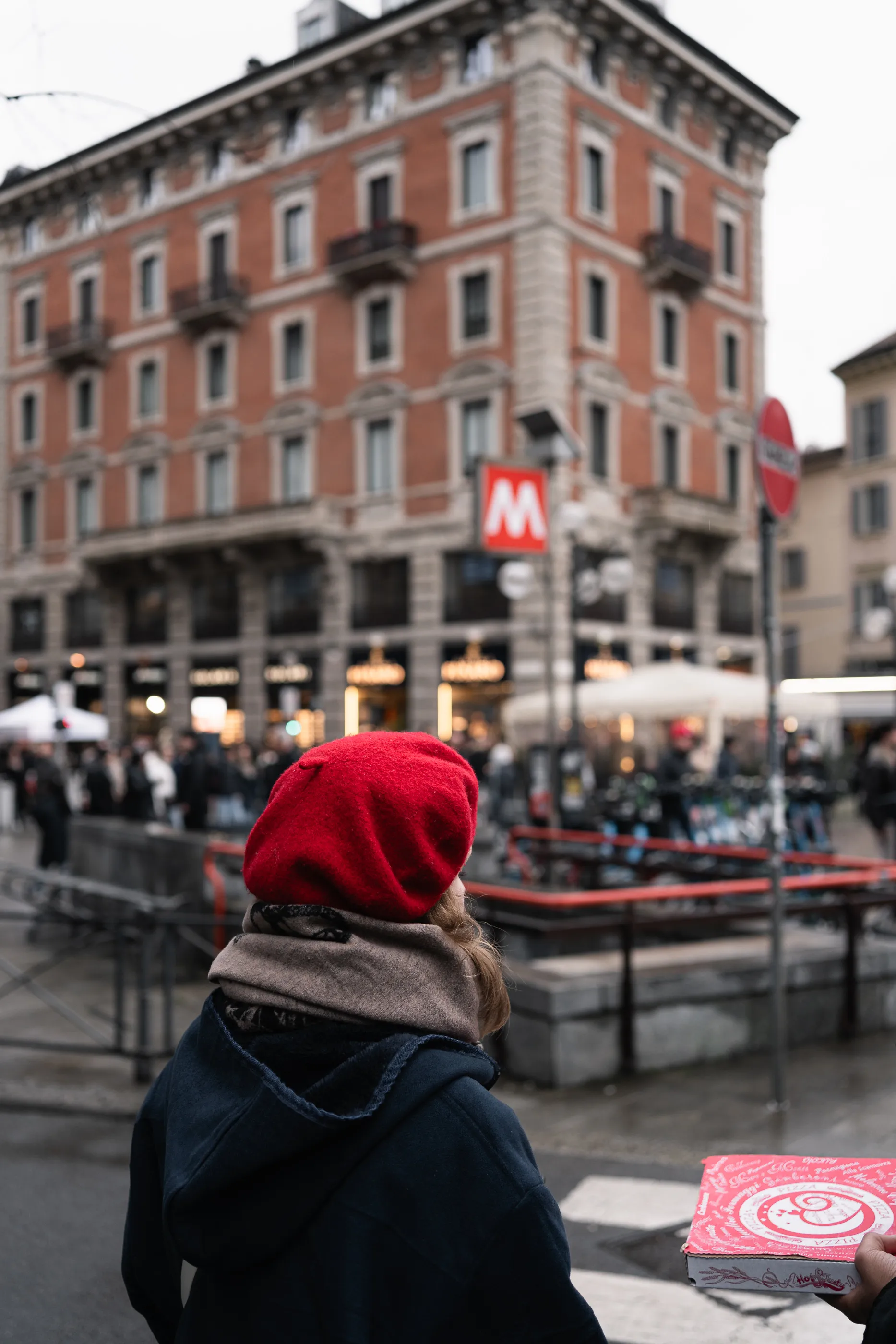 Seen from behind, a person in a red beret holds a pizza box near a Milan metro station.