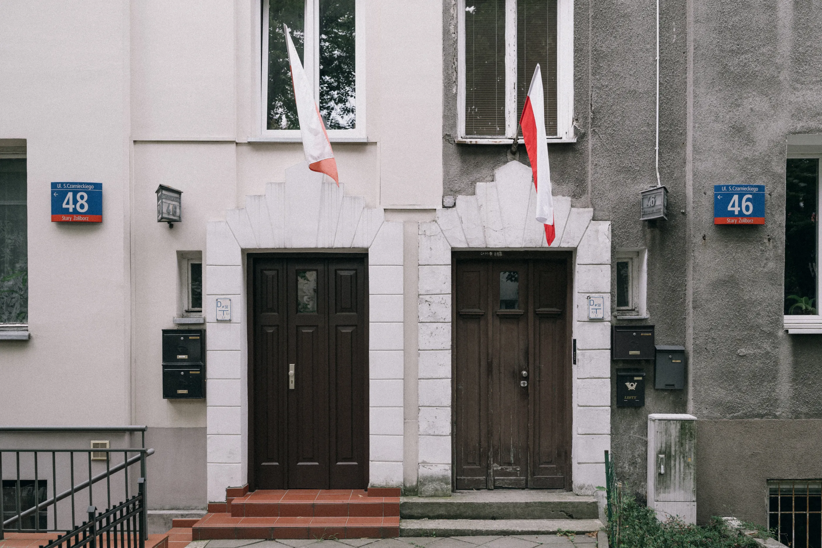 Two neighboring entrance doors on a Żoliborz street, each hung with a small Polish flag.